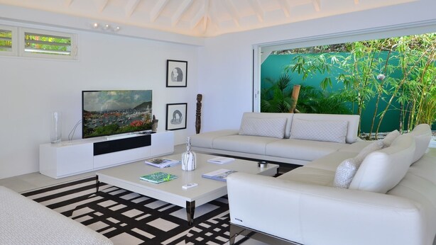 Een moderne woonkamer met een flatscreen televisie, een witte bank en een groot raam met uitzicht op groen.
