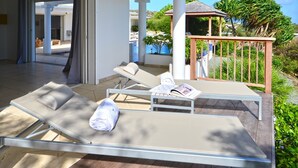 Ett soligt utomhusområde med en lounge, två solstolar, ett trädäck och utsikt över en pool och grönområden.