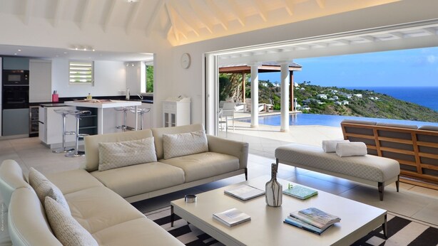 A modern living room with a sofa, coffee table, and ottoman, a kitchen area, and a large glass door leading to a pool and ocean view. - Belombre Luxury villa (Marigot)
