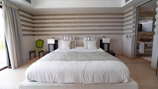 Un grand lit bien fait, avec une tête de lit rayée de blanc et de gris, deux tables de chevet avec des lampes, et une vue sur une salle de bain par une porte ouverte.
