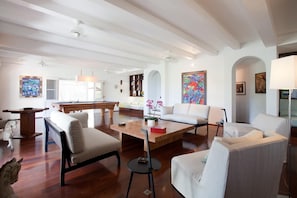 Amplia sala de estar con mesa de billar, sofás y una mesa de centro. Se observa una cocina al fondo y un comedor con techos altos y vigas a la vista.