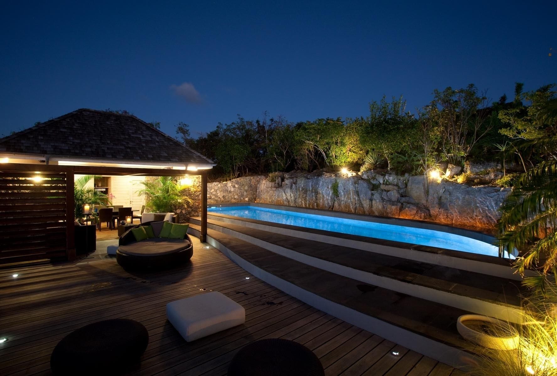 Un área junto a la piscina con un muelle de madera, un espacio de descanso cubierto y un muro de piedra.