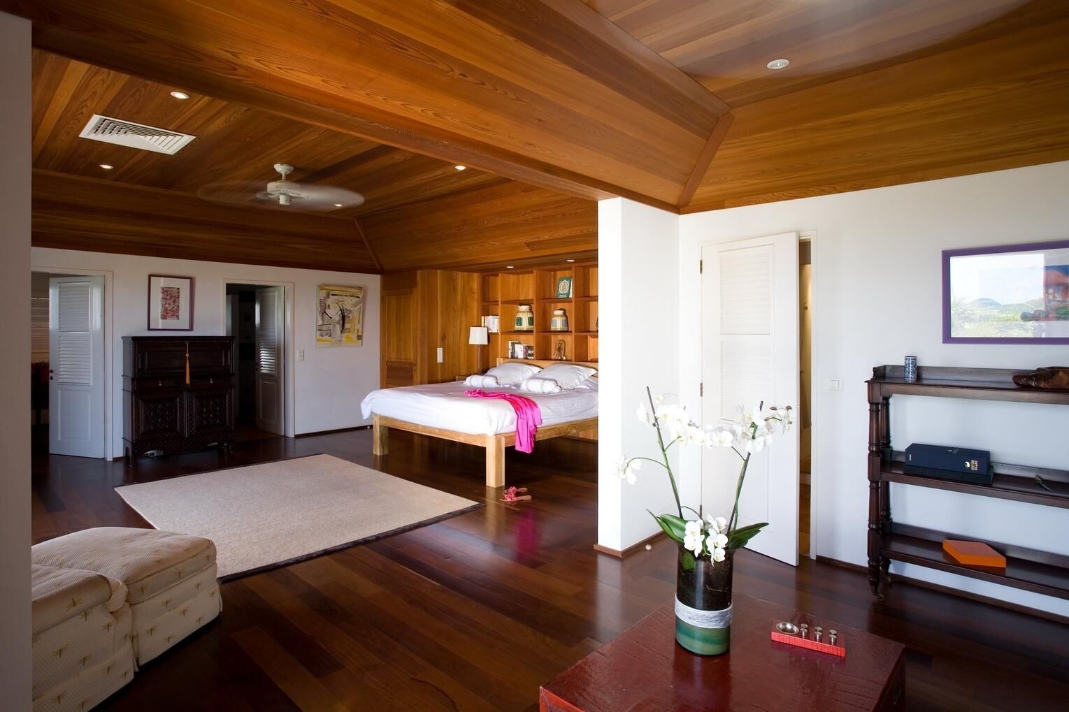 A spacious room with a large bed, wooden ceiling, and a wooden floor. There is a desk with a vase of flowers and a shelf with items.
