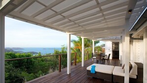 Een overdekt terras buiten met een houten vloer, een zithoek met een bank en stoelen, en uitzicht op de oceaan en heuvels.