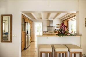A modern kitchen with wooden cabinets, a stainless steel refrigerator, a gas stove, and a large window with a view. There are three wooden stools in front of the counter.