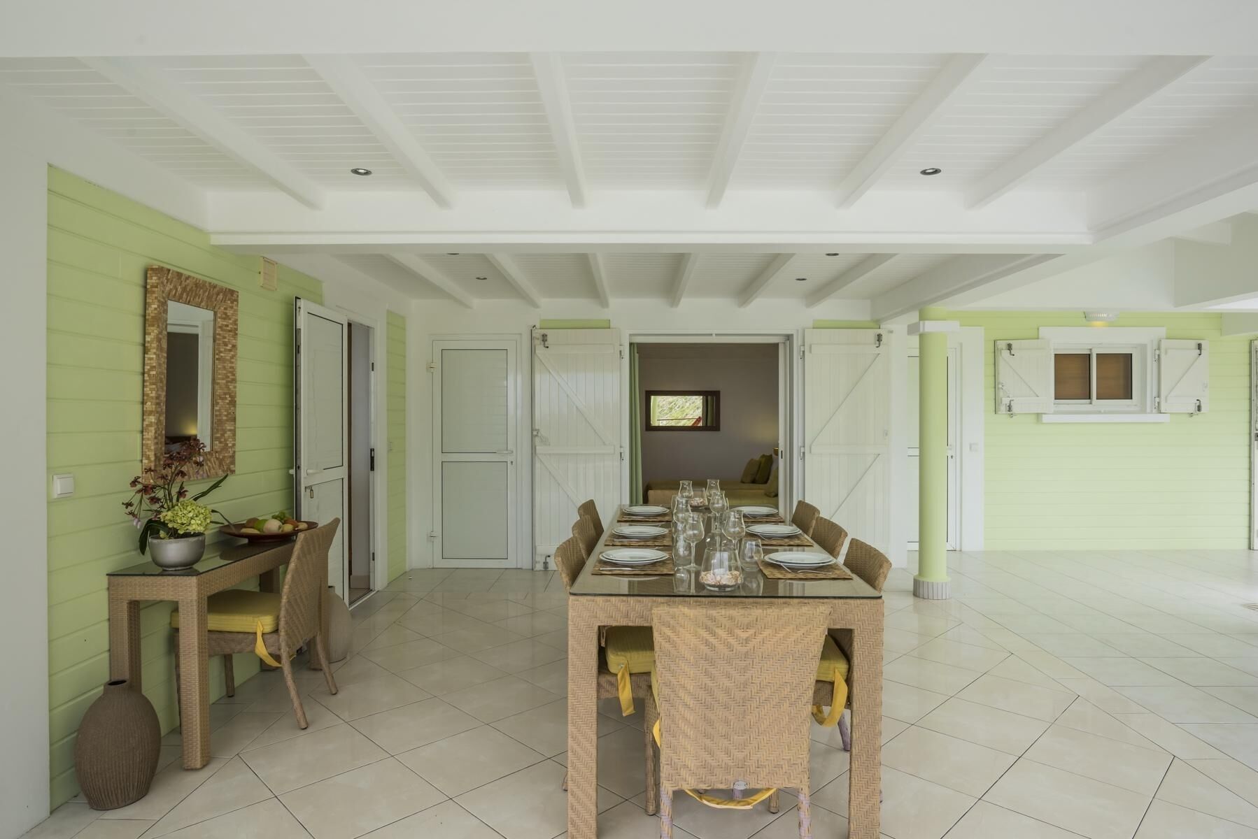 Zona de menjador amb una taula llarga preparada per a sis, envoltada de cadires, ubicada en una habitació amb parets verdes i bigues de sostre blanques.