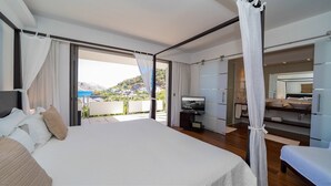 A modern hotel room with a large bed, a television, a bathroom with a large mirror, and a balcony with a view of the sea and hills. - Matajagui Luxury villa (Flamands)