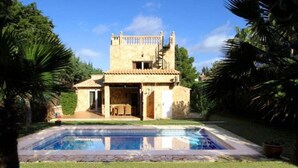 A two-story stone building with a tiled roof, a swimming pool in the foreground, and lush greenery surrounding the property. - Villa de Bonaire, Alcudia, large garden + pool, close to the beach, WiFi, for 8 people (Alcudia/Bon Aire)