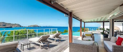 A covered outdoor deck with lounge chairs and a wicker sofa, overlooking a clear blue sea and distant hills.