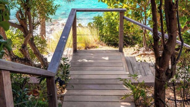 Eine Holztreppe führt zu einem Strand mit freiem Blick auf den Ozean und einer bergigen Landschaft im Hintergrund.