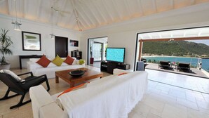 Ein modernes Wohnzimmer mit einem weißen Sofa, roten und schwarzen Kissen, einem Holztisch und einem Fernseher. Ein Balkon bietet Blick auf das Meer und einen Berg.