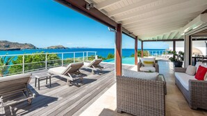 Eine überdachte Terrasse im Freien mit Liegestühlen und einem Rattansofa, mit Blick auf das klare, blaue Meer und sanfte Hügel in der Ferne.