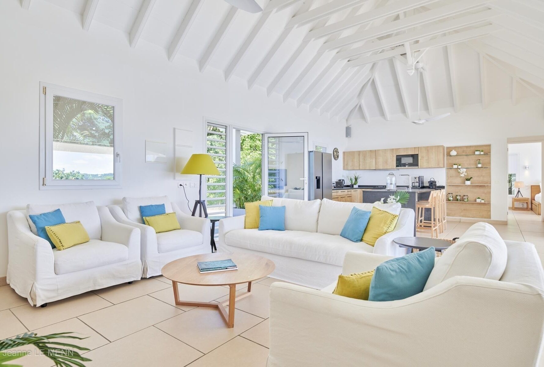 Un salon spacieux avec des canapés blancs, une table basse en bois et une vue sur la cuisine et l’espace extérieur.