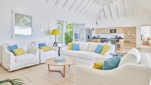 Een ruime woonkamer met witte banken, een houten salontafel en uitzicht op een keuken en buitenruimte.