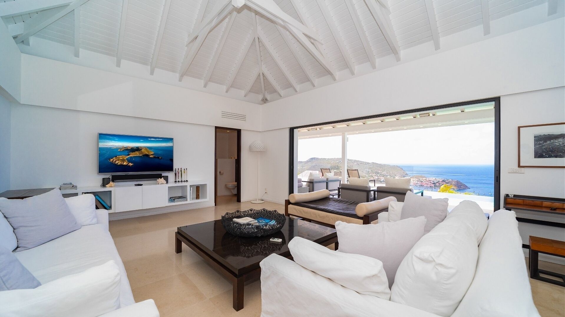 Un salon spacieux avec une grande télévision, des meubles blancs et une vue sur la mer.