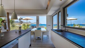 Une cuisine moderne avec vue sur l’océan, comprenant un coin repas avec des chaises et une table blanches, ainsi que de grandes fenêtres.