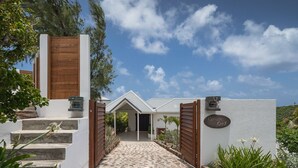A modern building with a stone staircase leading to a gated entrance, a sign reading "Captain Cook," and a clear blue sky with clouds. - Captain Cook (1/4 Rooms) (Pointe Milou)