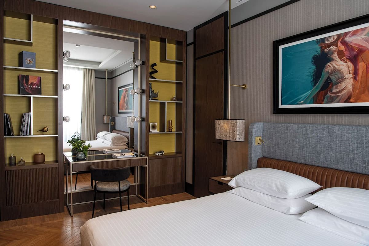 Een hotelkamer met een bed, een bureau, een stoel, een boekenplank en een schilderij aan de muur.