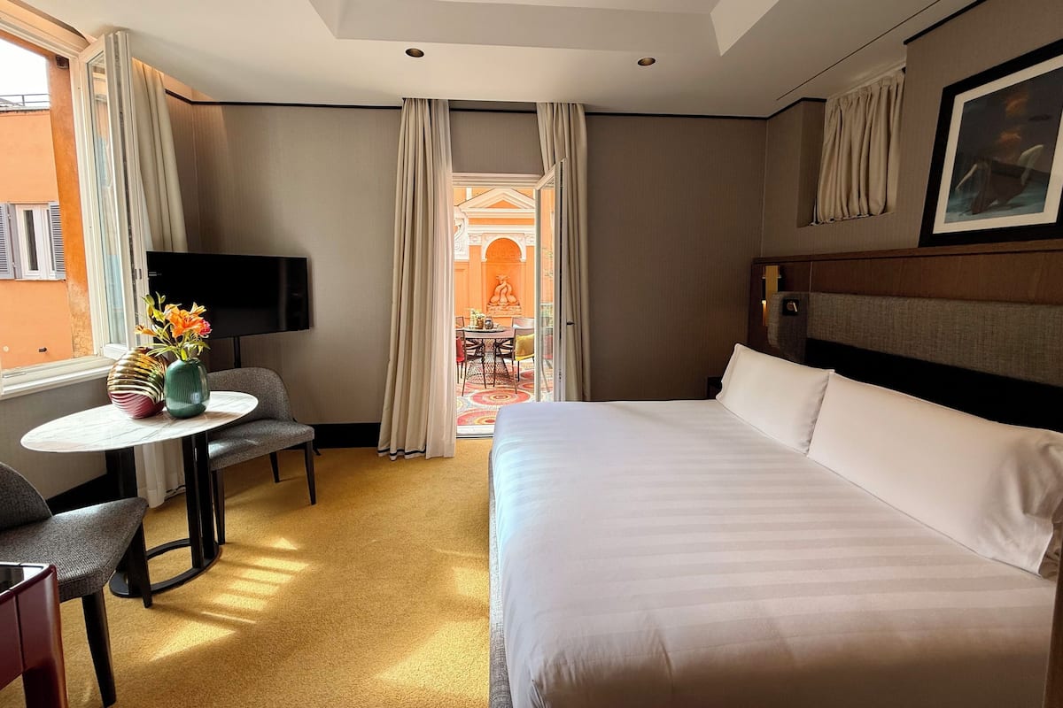 Een hotelkamer met een bed, een televisie, een kleine tafel met een vaas, en uitzicht op het balkon.