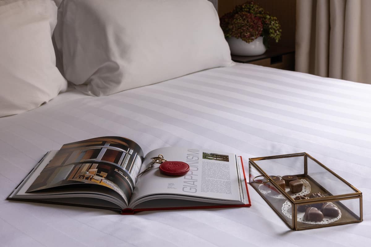 Een keurig opgemaakt bed met een boek, een schotel chocolade, een lamp en een ingelijst beeld aan de muur.
