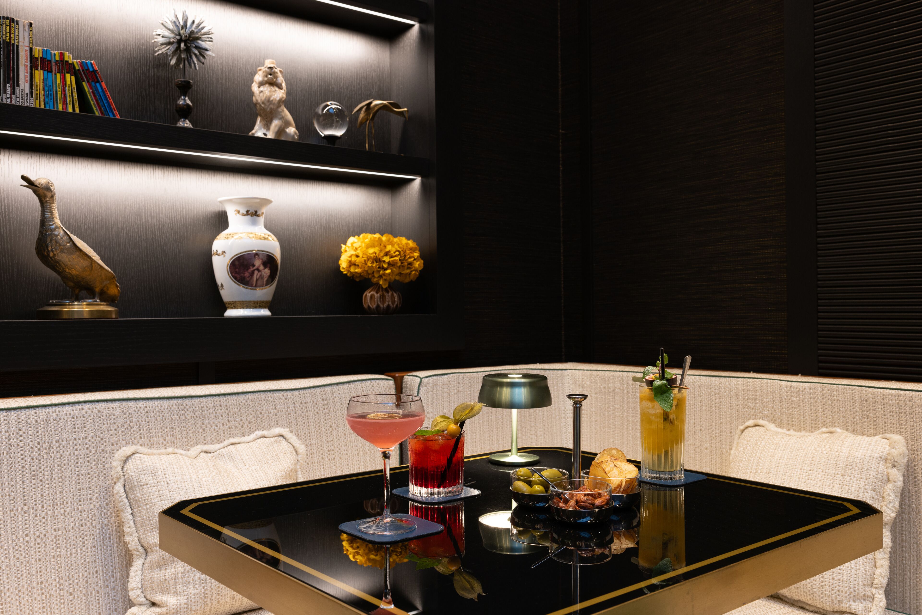 Un comedor con una mesa negra, sillas blancas con cojines, una estantería con objetos decorativos y una variedad de bebidas y aperitivos.