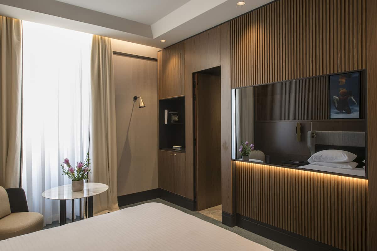 Een moderne hotelkamer met een groot bed, een klein rond tafeltje met een bloemenvaas, een ingebouwde televisie en een wandpaneel van hout.