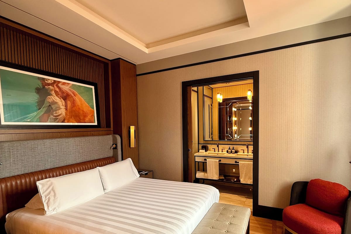 Een hotelkamer met een groot bed, een bankje, een rode stoel, een schilderij aan de muur en een spiegel in de hoek.