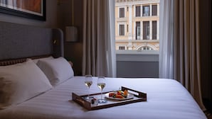 A neatly made bed with white linens, two glasses of white wine, and a tray with fruit and a small bottle on a wooden tray.