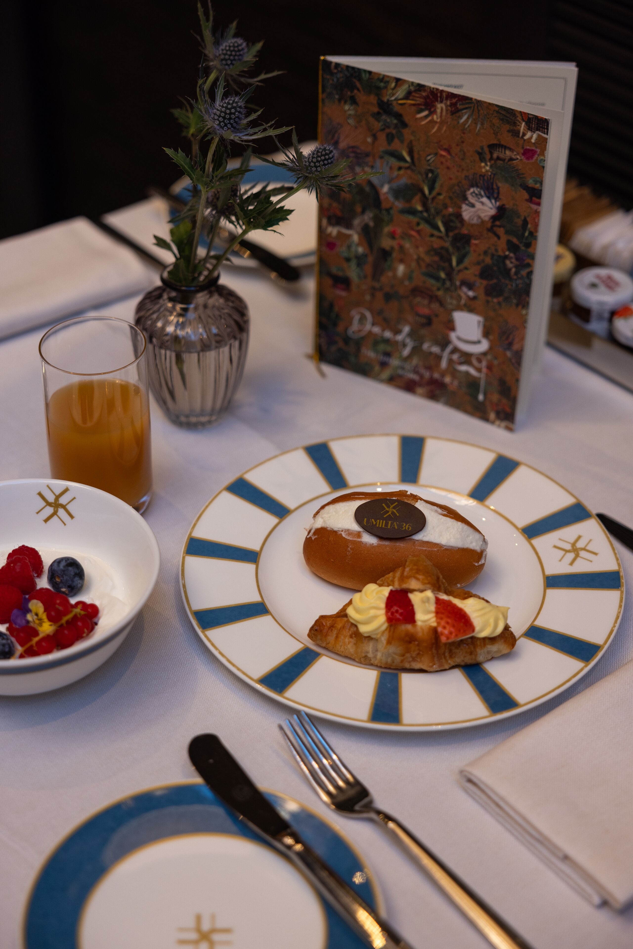 A table set with plates, a glass of orange juice, a vase with flowers, and a menu.