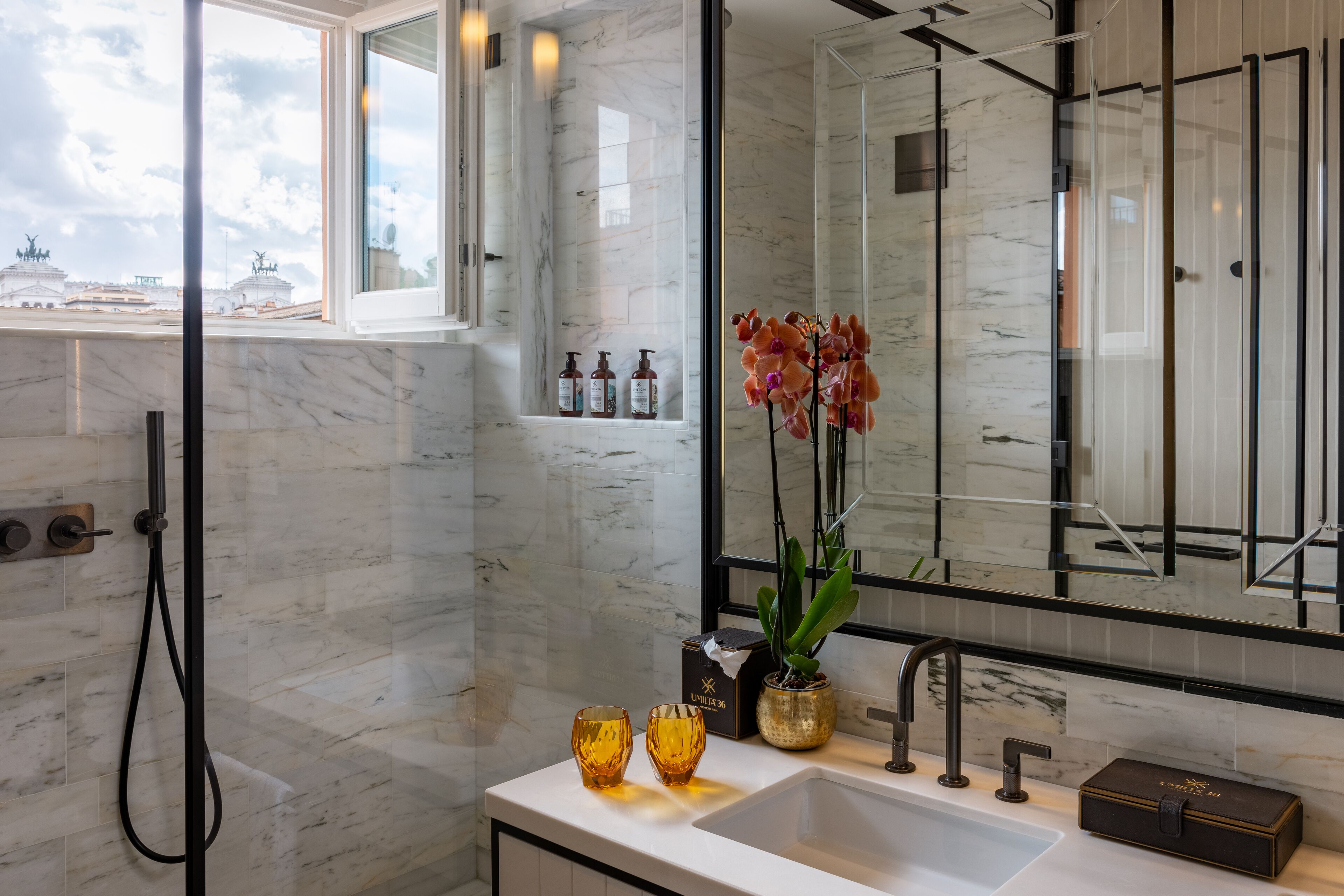 Un baño moderno con un espejo grande, una ducha y un lavamanos. Hay dos botellas de champú y acondicionador sobre la encimera, y un florero con flores.