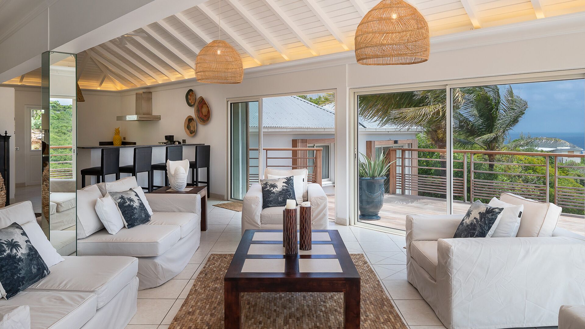 A modern living room with white sofas, a coffee table, and a large sliding glass door leading to a balcony with a view of the ocean.
