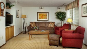 Ein Wohnzimmer mit einer Couch, Sesseln, einem Couchtisch, einem Fernseher und einer Lampe.