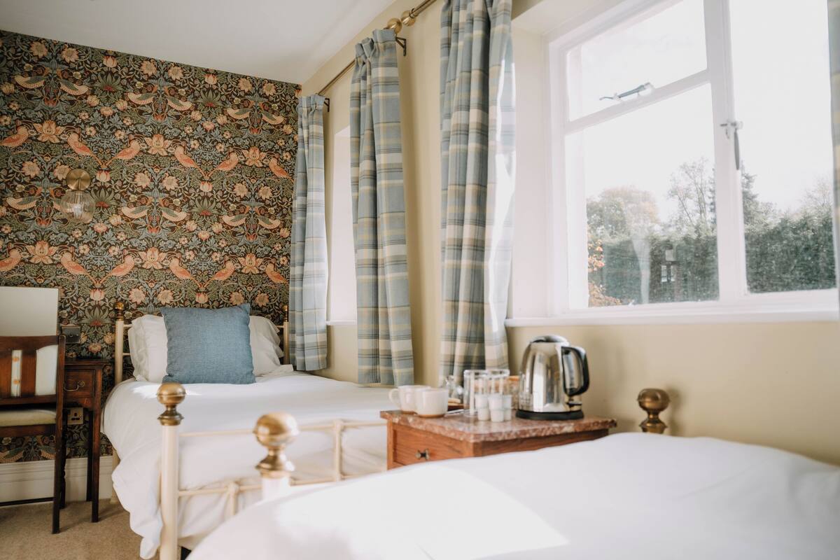 A bedroom with two beds, a wooden nightstand, a kettle, and a window with curtains.