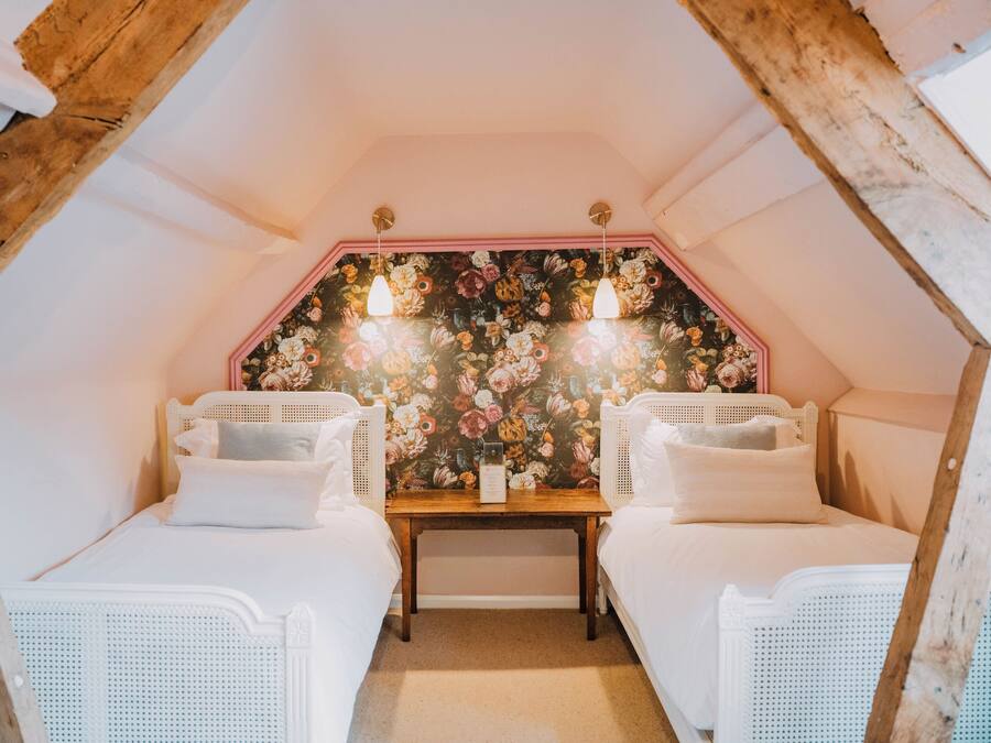 A cozy attic bedroom with two white wicker beds, a floral wallpaper, and a wooden ceiling.
