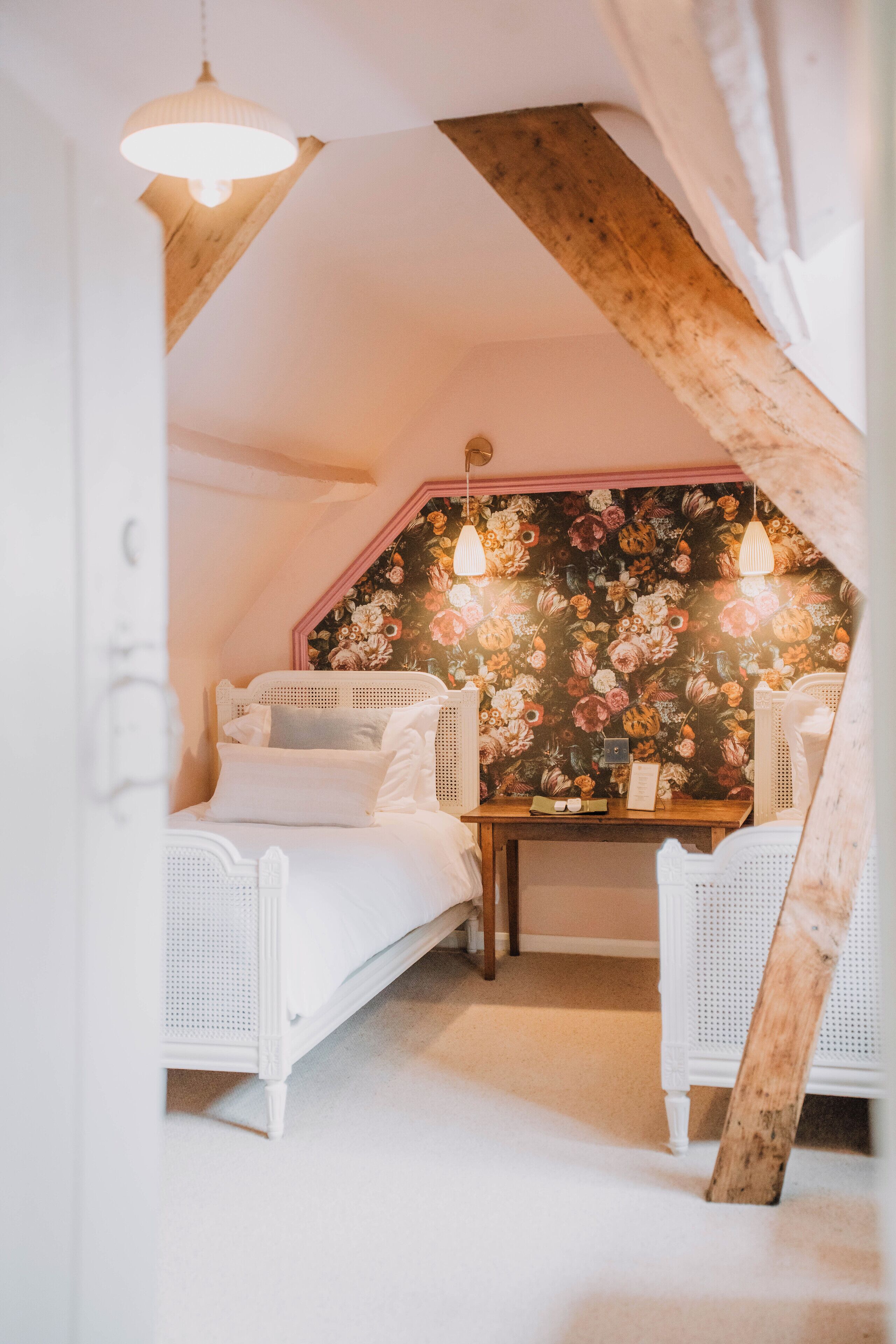Un dormitorio estilo ático acogedor, con papel tapiz floral, una cama blanca y una mesa de madera.