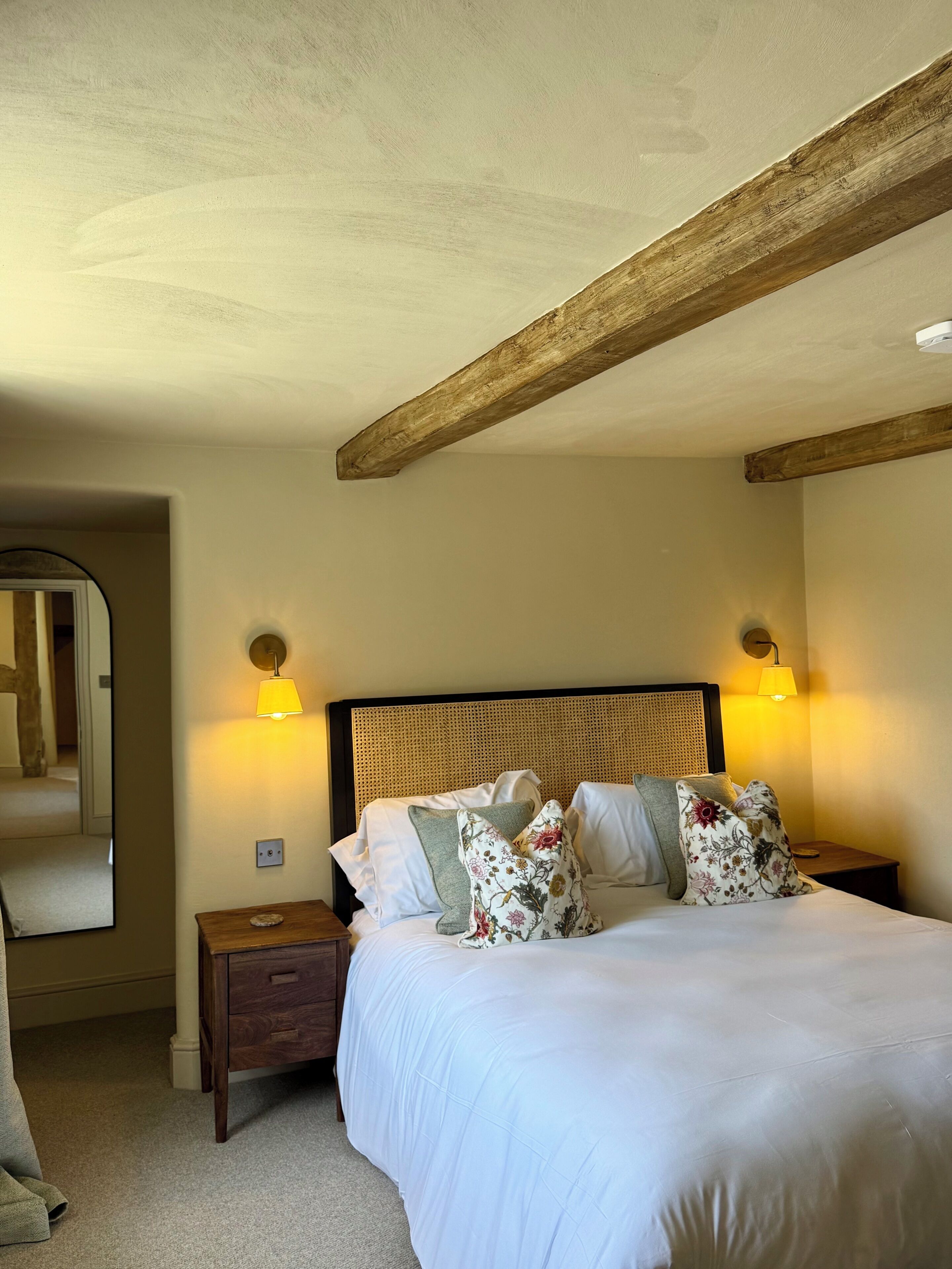 Une chambre avec une poutre en bois au plafond, un lit avec une literie blanche et des coussins à motifs floraux, deux lampes de chevet, une table de chevet en bois et un miroir sur le mur.
