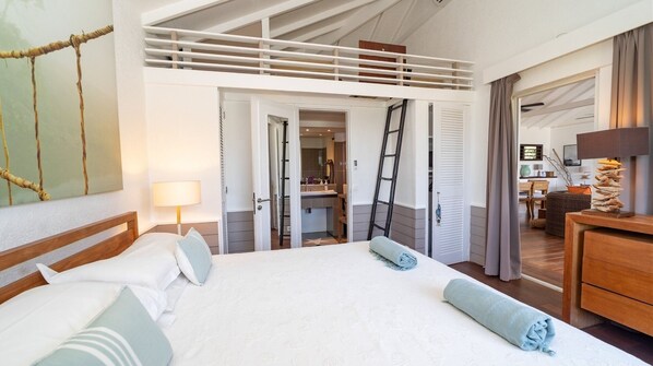 A spacious bedroom with a large bed, a lofted sleeping area, a bathroom with a mirror, and a view of the living area.