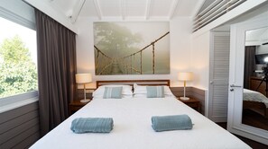 Una habitación de hotel con una cama grande, dos mesitas de noche con lámparas, un cuadro de un puente y una puerta que da a un balcón.