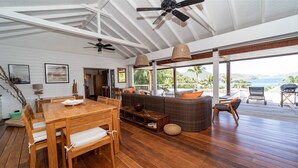 Une pièce spacieuse avec une table à manger en bois, un canapé et un ventilateur de plafond. De grandes fenêtres offrent une vue sur l’océan.