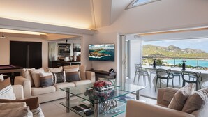 A modern living room with a glass coffee table, a sofa with patterned cushions, a kitchen area with appliances, and a large TV. - Panama (3/5 Rooms) Luxury villa (Saint Jean)