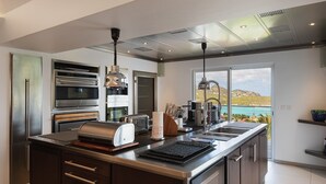 A modern kitchen with built-in appliances, a large island, and a view of a coastal landscape. - Panama (3/5 Rooms) Luxury villa (Saint Jean)