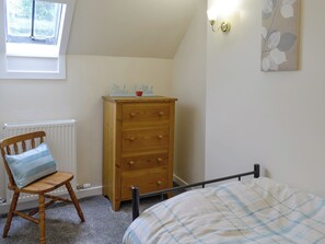 Une chambre à coucher comprenant un lit, une chaise en bois avec un coussin, une commode en bois, un radiateur et un luminaire fixé au mur.
