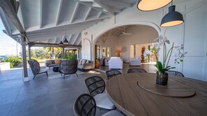Une vaste terrasse dotée d’une table et de chaises en bois, un espace salon couvert avec des canapés et des fauteuils, et une vue sur l’extérieur.