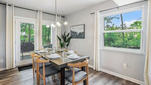 Un espace repas comprenant une table en bois, des chaises à revêtement gris, un luminaire suspendu moderne et de grandes fenêtres donnant sur un paysage verdoyant.