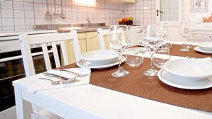 Una mesa de comedor dispuesta con platos, fuentes y copas de vino blancos, rodeada de sillas blancas en una cocina con armarios y salpicadero de azulejos.