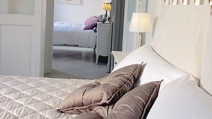 Una cama bien tendida con cubrecama acolchado y dos almohadas, una mesita de noche con lámpara, y una puerta que da a otra habitación.
