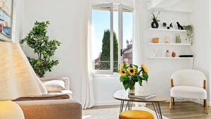 A modern living room with a sofa, armchair, and ottoman, a small table with a vase of sunflowers, and a window with a view of a building and trees. - 04. Bright apartment with balcony and sea view, close to the beach (Saint-Jean-Cap-Ferrat)