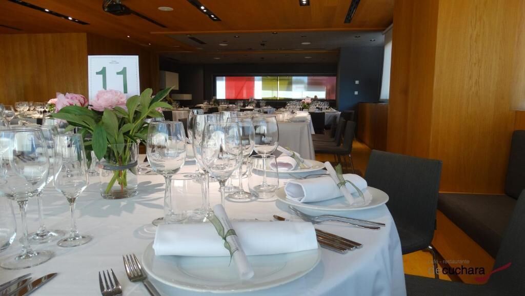 Un salón de banquetes con mesas redondas dispuestas para un evento formal, cada una con manteles blancos, platos, cubiertos y copas de vino.
