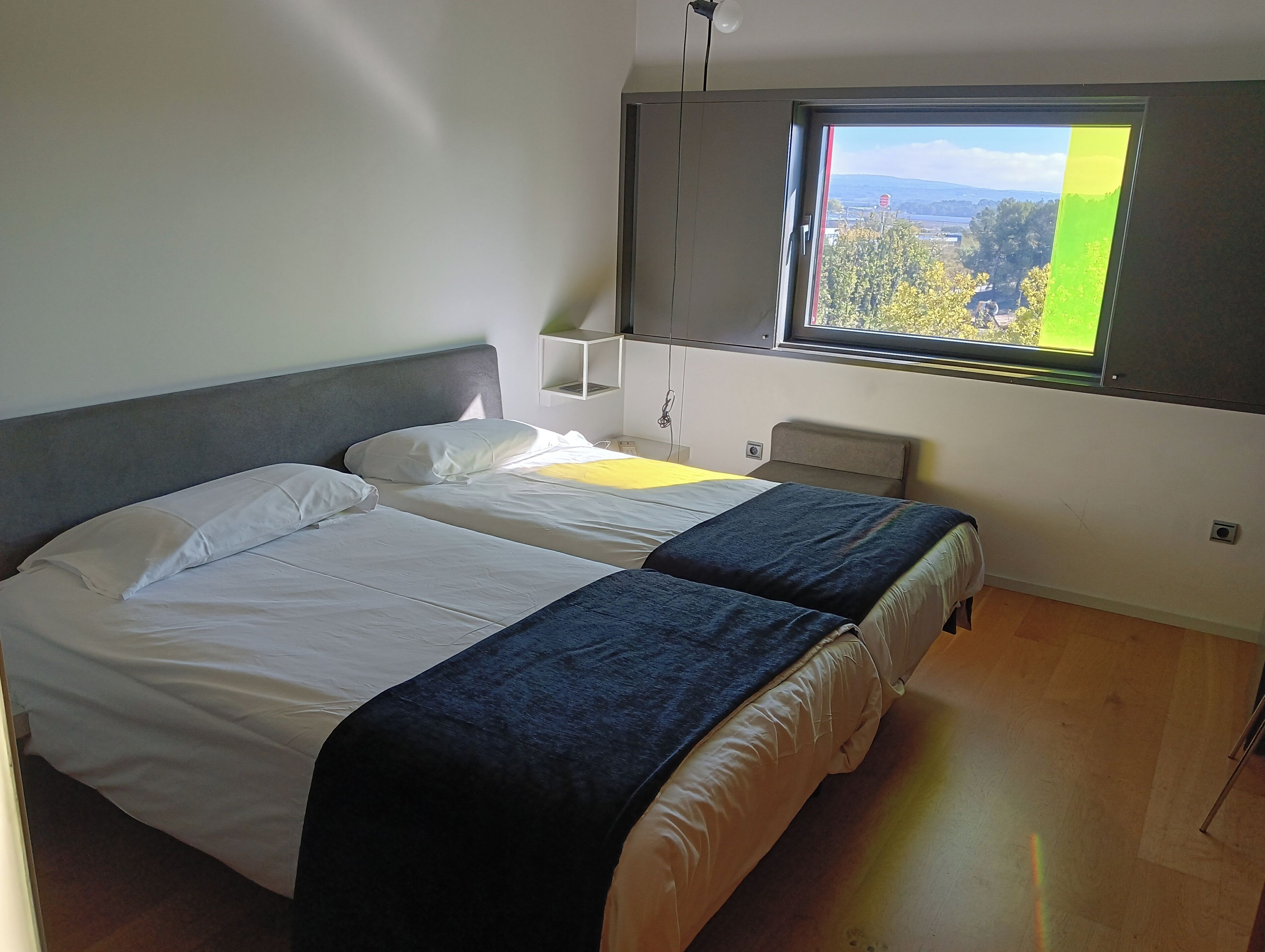 Un dormitorio con dos camas, una ventana que da a zonas verdes, una mesita de noche blanca y un estante montado en la pared.