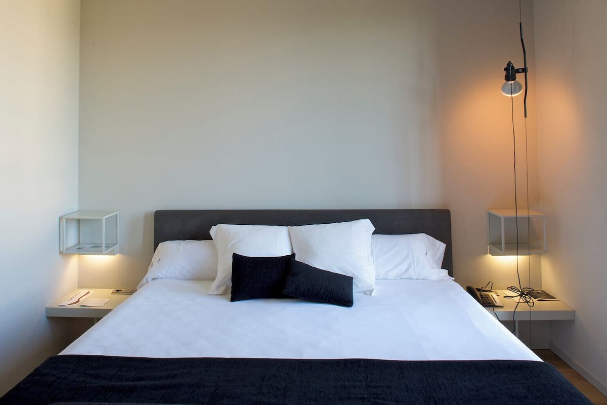 Een net opgemaakt bed met witte en zwarte kussens, twee nachtkastjes met lampen en een telefoon op het nachtkastje.